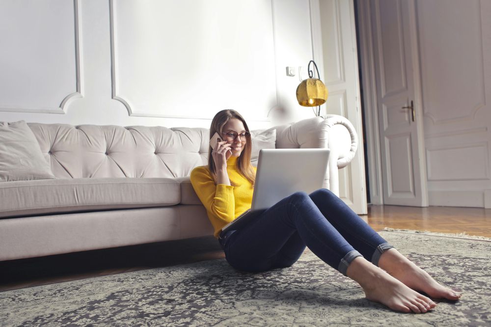 Eine lächelnde Frau sitzt vor Ihrer Couch und arbeitet an Ihrem Laptop während Sie telefoniert